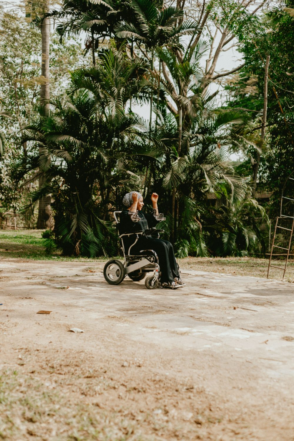 A woman in a wheelchair smiling and celebrating with arms raised in a leafy garden