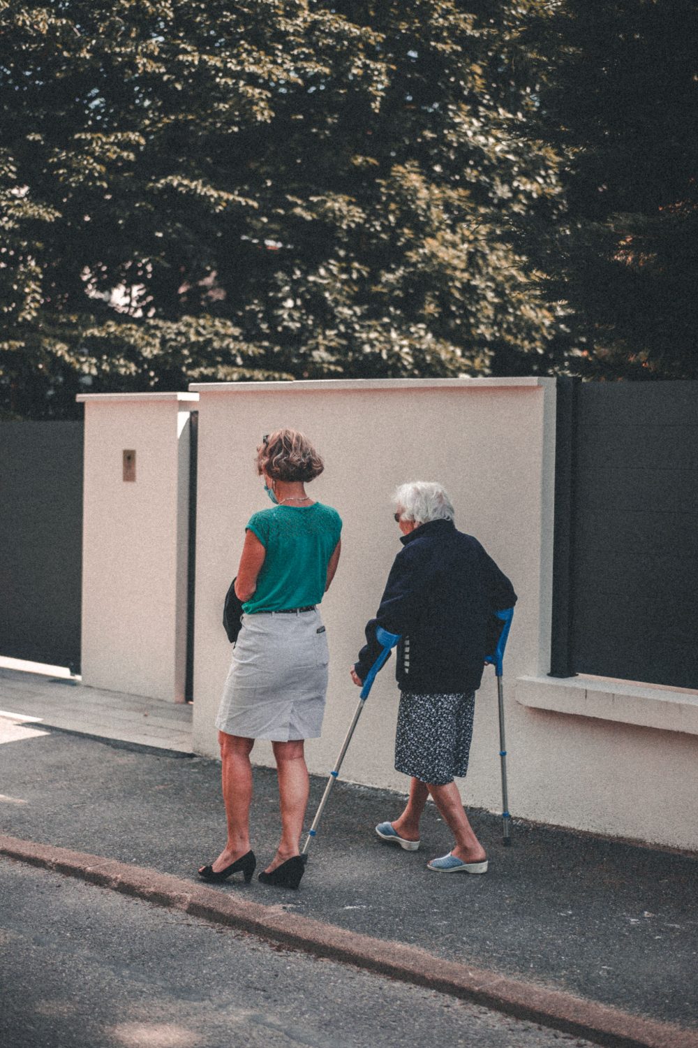A support worker walking alongside an older woman using crutches, outdoors